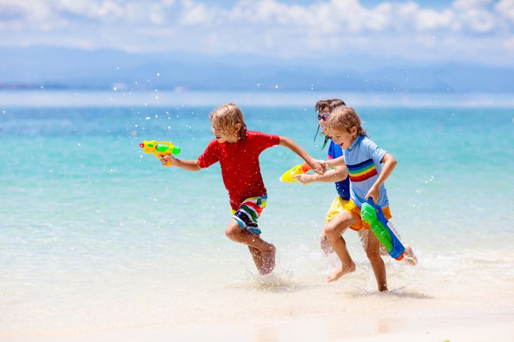 Drei Kinder rennen lachend mit bunten Wasserpistolen am Strand entlang, während Meereswellen an den hellen Sand rollen und sommerliches Sonnenlicht die Szene erhellt