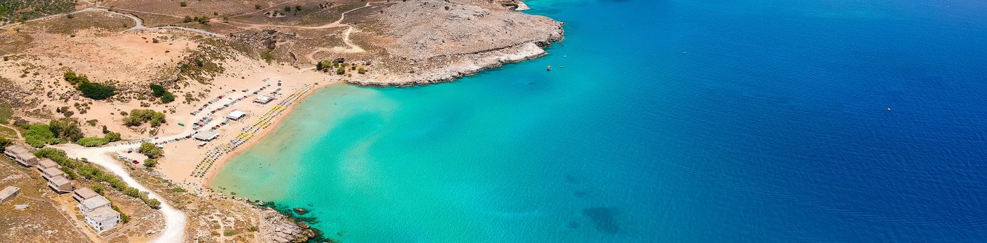 Luftaufnahme eines Strandes mit türkisfarbenem Wasser, felsiger Küste und trockenem, hügeligem Hinterland.