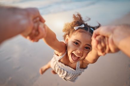 Mädchen wird am Strand von einem Erwachsenen spielerisch an den Händen im Kreis geschwungen, während die Sonne warm auf den Sand und das Meer scheint