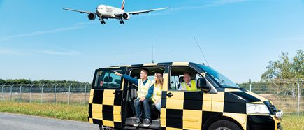 Vorfeldrundfahrt am Flughafen Hamburg: Besucher sitzen in einem gelb-schwarzen Shuttlefahrzeug und beobachten ein landendes Passagierflugzeug über dem Rollfeld