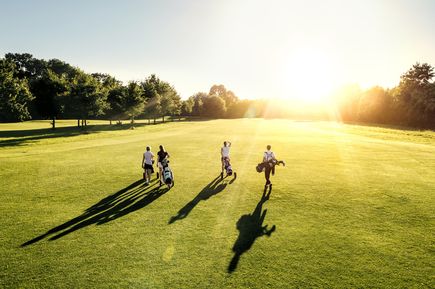 Gruppe von Personen geht mit Golfschlägern über einen sonnigen Golfplatz, lange Schatten auf dem grünen Rasen, Bäume im Hintergrund und strahlende Sonne am Horizont