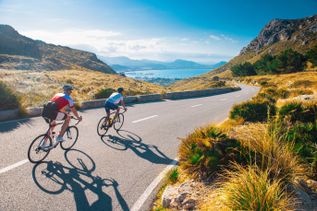 Zwei Radfahrer auf einer kurvigen Bergstraße mit Blick auf das Meer und die Bucht, unter blauem Himmel
