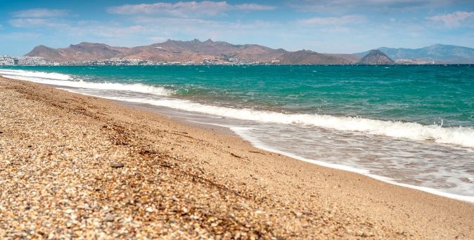 Strand mit Kieselsteinen und Wellen am Ufer vor einer Berglandschaft unter blauem Himmel an der Küste von Kos