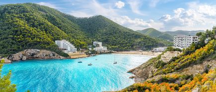Blick auf die Bucht Cala Llonga auf Ibiza mit türkisblauem Meer, bewaldeten Hügeln, weißen Hotelgebäuden am Strand und Booten im Wasser