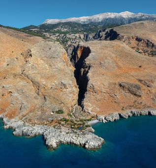 Felsige Küstenlandschaft mit schmaler Schlucht, die ins Meer mündet, unter blauem Himmel.