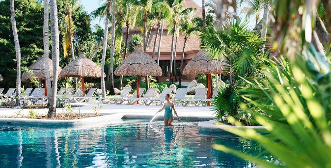 Person im Pool eines der Bahia Principe Hotels & Resorts, umgeben von tropischer Gartenlandschaft, Palmen, Sonnenliegen und strohgedeckten Sonnenschirmen