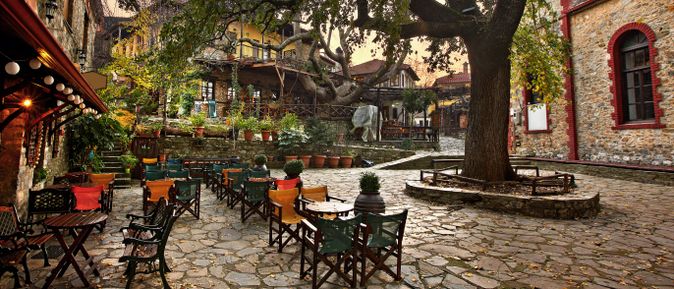 Innenhof eines Restaurants mit gepflastertem Boden, mehreren Tischen und Stühlen, großen Bäumen und historischen Gebäuden im Hintergrund.