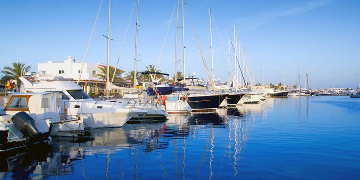 Hafen von La Savina auf Formentera mit weißen Gebäuden, Palmen und zahlreichen Segel- und Motorbooten, die sich im klaren Wasser spiegeln