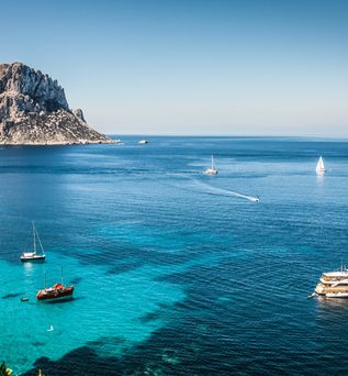 Blick auf das klare blaue Meer bei Ibiza mit mehreren Segelbooten und einer markanten Felseninsel im Hintergrund