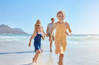 Zwei Kinder laufen lachend am Strand entlang, während die Eltern im Hintergrund Hand in Hand folgen, sonniger Tag am Meer