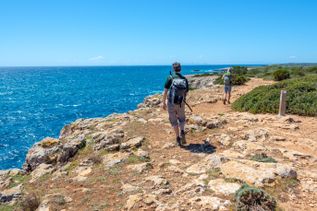 Zwei Personen wandern entlang eines felsigen Küstenpfads mit Blick auf das tiefblaue Meer und die weite Landschaft unter klarem Himmel