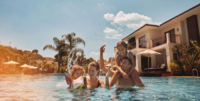 Familie mit zwei Kindern im Außenpool des Ela Excellence Resort Belek, vor eleganten Hotelgebäuden, Palmen und Wasserspielen in der weitläufigen Resortanlage.