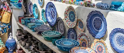 Regal mit handbemalten Keramiktellern und Schalen in Blau-, Weiß- und Orangetönen, darunter braune Töpferwaren.