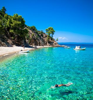 Klares türkisfarbenes Meer mit Schwimmer, felsige Küste mit Pinien und kleinen Booten im Wasser.