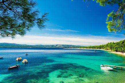 Türkisblaues Meer am Strand Zlatni Rat auf der Insel Brač in Kroatien mit kleinen Booten, bewaldeter Küste und Blick auf die dalmatinische Adriaküste