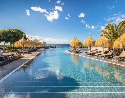 nfinity-Pool mit Stufen, umgeben von Liegestühlen und Strohdach-Sonnenschirmen, Palmen und blauem Himmel am Barceló Tenerife.