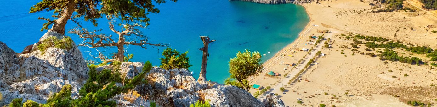 Luftaufnahme eines Strandes mit türkisfarbenem Wasser, felsiger Küste und trockenem, hügeligem Hinterland.