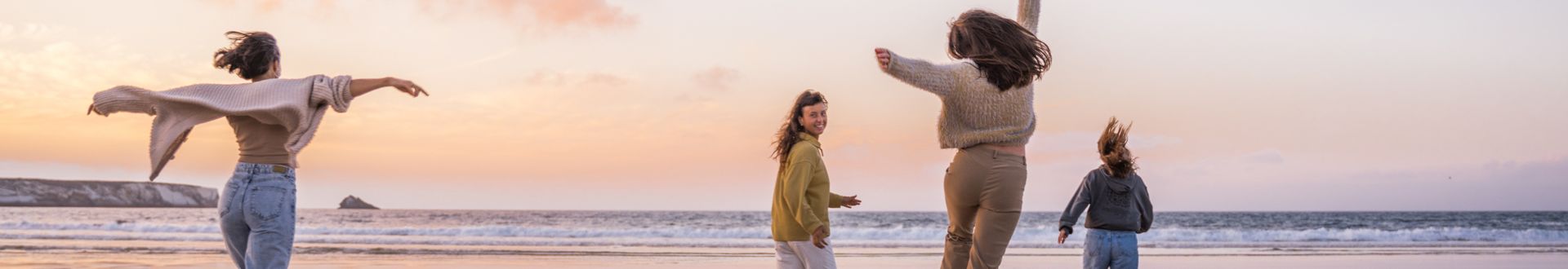 Vier Personen bewegen sich dynamisch am Strand entlang, während die Sonne in warmen Farben über dem Meer untergeht; Weitwinkelaufnahme mit ausgebreiteten Armen und springenden Silhouetten vor einer ruhigen Küstenlandschaft