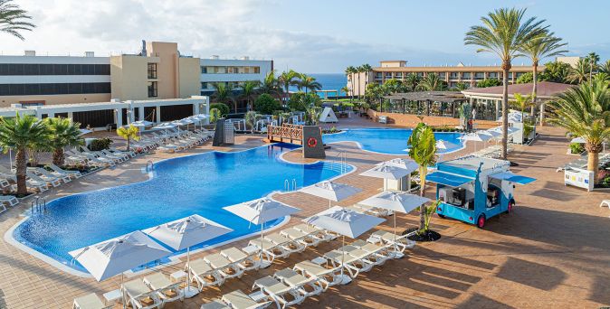 Großer Poolbereich des Iberostar Waves Gaviotas Park mit Liegen, Sonnenschirmen, Palmen und einem kleinen Verkaufswagen auf der Terrasse.